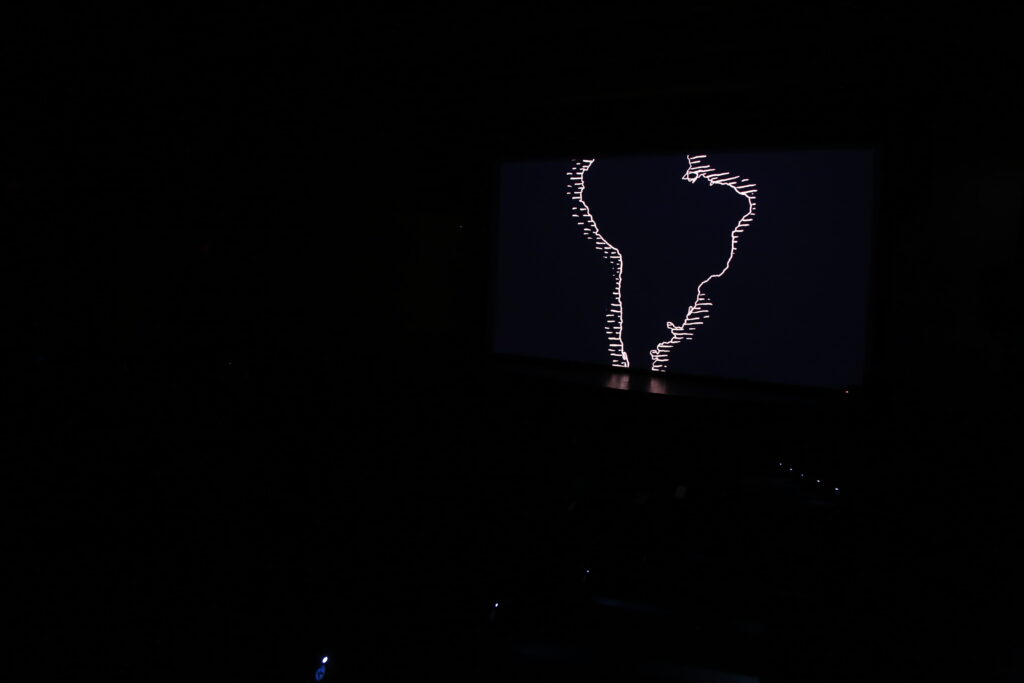 Sala de cinema escura. Na tela se vê uma ilustração representando o contorno da America do Sul.