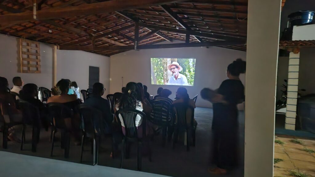 Sala escura. Pessoas assistem sentadas à projeção de um filme.
Na tela vemos um senhor de óculos e chapéu de palha falando.